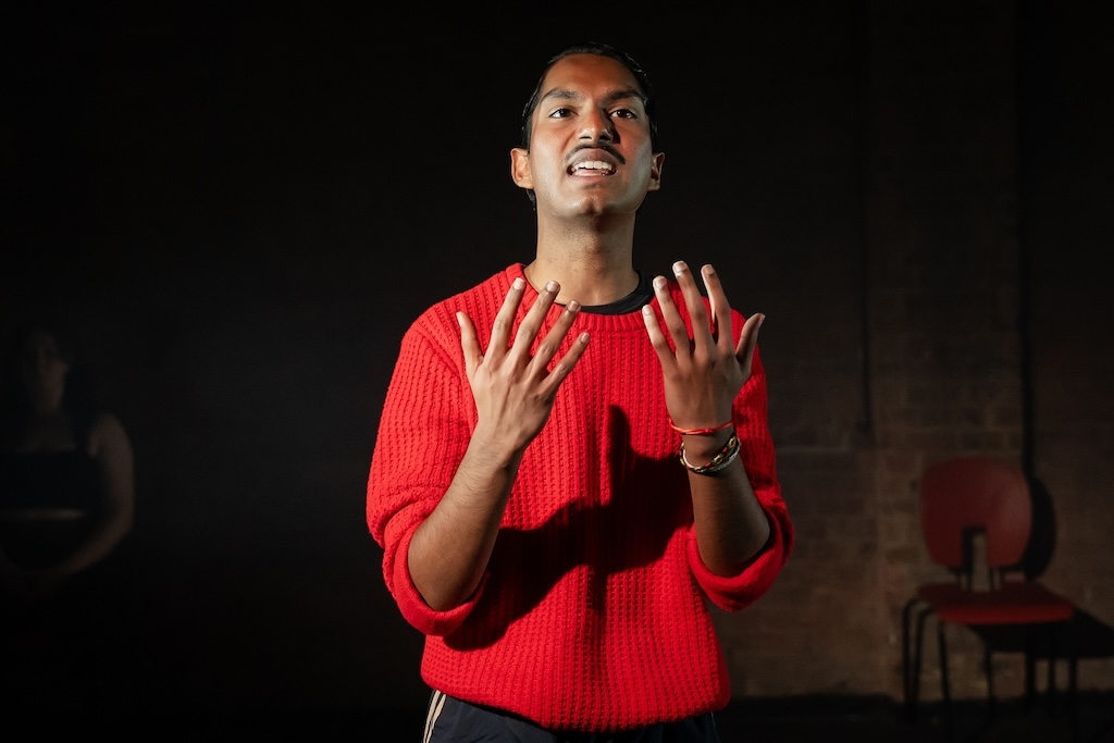 Action shot of the spring 2024 Shakespeare in Performance at RADA final presentation of Julius Caesar. An actor wearing a bright red sweater holds his hands in front of his chest in mid-performance.